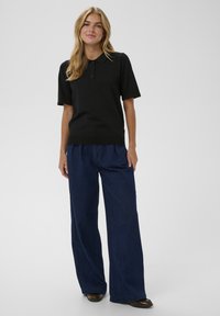 Camisa polo negra de manga corta, con dobladillo acanalado y tapeta de tres botones, combinada con pantalones de mezclilla oscuros de pierna ancha y zapatos marrones.