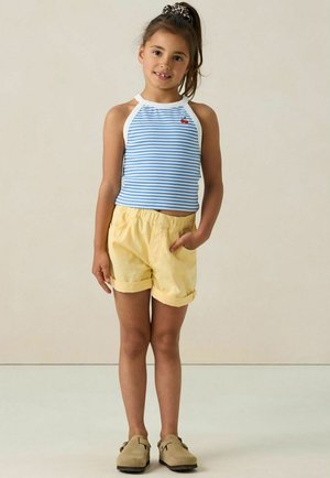 Haut à col halter rayé bleu clair et blanc avec un petit graphique de cerise rouge, associé à un short jaune et des sabots beige. Textures lisses, coupe décontractée.