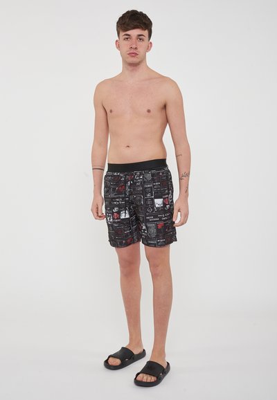 Pantaloni da bagno neri con una stampa grafica che presenta vari motivi in bianco e rosso. Vita elastica. Modello in piedi a piedi nudi con sandali.