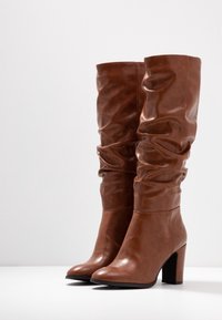 Botas de rodilla de imitación cuero marrón con un diseño arrugado, puntera redonda y tacón cuadrado negro. Textura suave con detalles fruncidos.