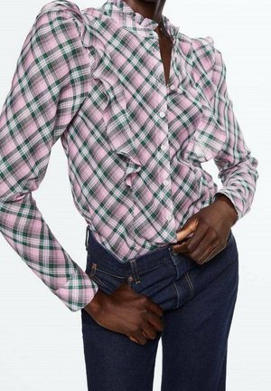 Osoba o ciemnej karnacji ubrana w różowo-zieloną kratowaną bluzkę z falbanami oraz ciemnoniebieskie dżinsy, regulująca pas obiema rękami.