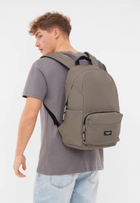 Grå ryggsäck med vadderade axelremmar, framficka och logodetalj. Slät, hållbar tyg med en strukturerad form.