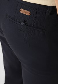 Granatowe spodnie z fakturowanej tkaniny z tylną kieszenią, która ma niebieski guzik oraz skórzaną naszywkę z logo "bugatti."