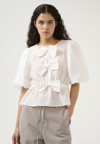 Weiße Bluse mit Puffärmeln, drei auffälligen Schleifenakzenten, taillierter Taille und strukturiertem Stoff. Kombiniert mit hellgrauen Hosen mit Nieten-Details.
