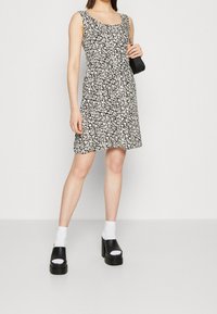 Nainen, joka on pukeutunut ilman hihoja musta-valkoiseen kukkamekkoon, valkoisiin sukkiin, mustiin korkokenkiin ja pitää pientä mustaa käsilaukkua.