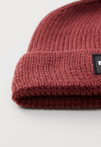 Folded knitted maroon beanie hat with a visible black and white rectangular label on a white surface.