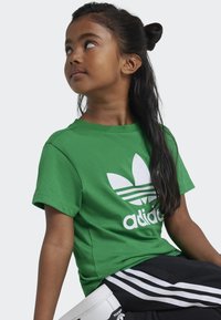 Zelené bavlněné tričko s velkým bílým logem adidas, které je doplněno designem listu. Krátké rukávy a kulatý výstřih.