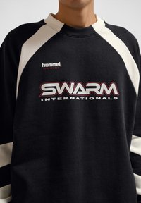 Close-up of a person wearing a black and white sweatshirt with "hummel sportswear" and "SWARM INTERNATIONALS" logos on the front.