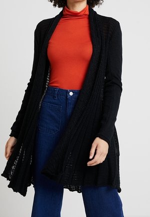 Personne portant un cardigan noir en maille ouvert sur un col roulé orange et un jean bleu taille haute avec une poche avant.