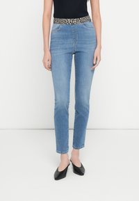 Sinised teksapüksid, millel on leopardimustriline vöökoht, kitsas lõige ja standardne viiepaneeliline taskute disain. Sobitatud mustade libisevate kingadega.