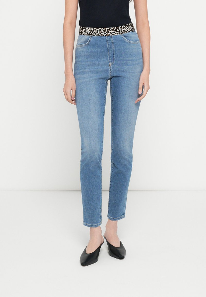 Sinised teksapüksid, millel on leopardimustriline vöökoht, kitsas lõige ja standardne viiepaneeliline taskute disain. Sobitatud mustade libisevate kingadega.