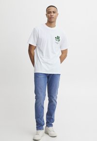  camiseta blanca de algodón con un gráfico floral verde en el pecho, combinada con jeans azul claro y zapatillas blancas. Diseño simple.