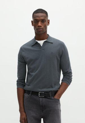 Uomo che indossa una camicia grigia a maniche lunghe con colletto sopra una maglietta bianca, cintura nera e jeans grigio scuro, in piedi con una mano in tasca.