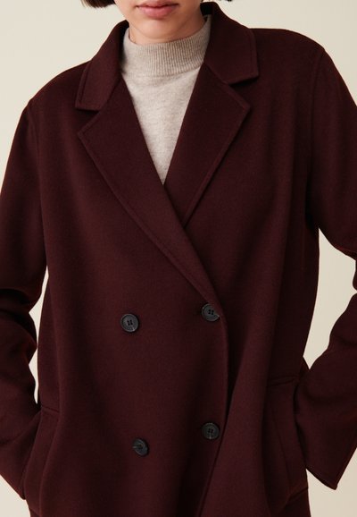 Cappotto bordeaux doppiopetto realizzato in morbido misto lana, con bottoni neri, rever a serratura e tasche laterali. Tessuto testurizzato in evidenza.
