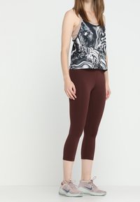 Kobieta w bezrękawnikowej bluzce w czarno-białe wzory, maroonowych legginsach do kostek i jasnoróżowych trampkach Nike, stojąca na jednolitym tle.