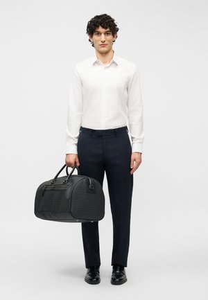 Man draagt een wit overhemd en donkere broek, houdt een zwarte reistas met patroon aan de handvatten vast, staat tegen een effen achtergrond.