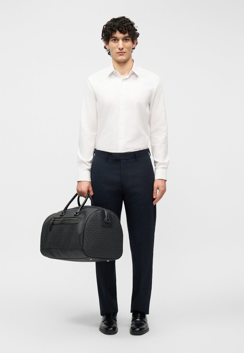 Άνδρας που φοράει λευκό πουκάμισο και σκούρο παντελόνι, κρατώντας μια μαύρη τσάντα ταξιδιού με σχέδια από τις λαβές, στέκεται μπροστά σε ένα απλό φόντο.