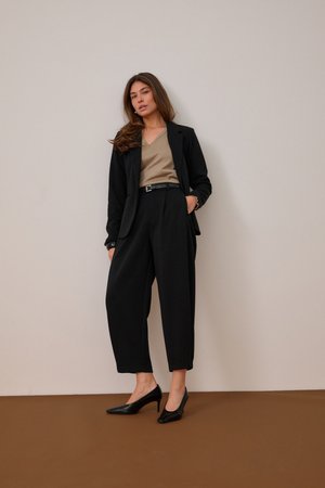Femme debout contre un mur beige, portant un blazer noir, un haut beige à col en V, un pantalon noir taille haute et large, ainsi que des talons noirs, une main dans la poche.