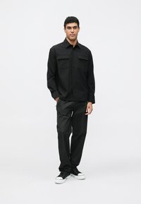 Cămașă neagră cu nasturi și două buzunare la piept, asortată cu pantaloni negri și adidași negri cu talpă albă. Design simplu, modern; textură netedă.