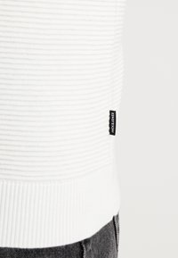 Vit ribbad tröja med texturerad design och svart etikettdetalj. Har en åtsittande passform som framhäver horisontella balkar längs kroppen.