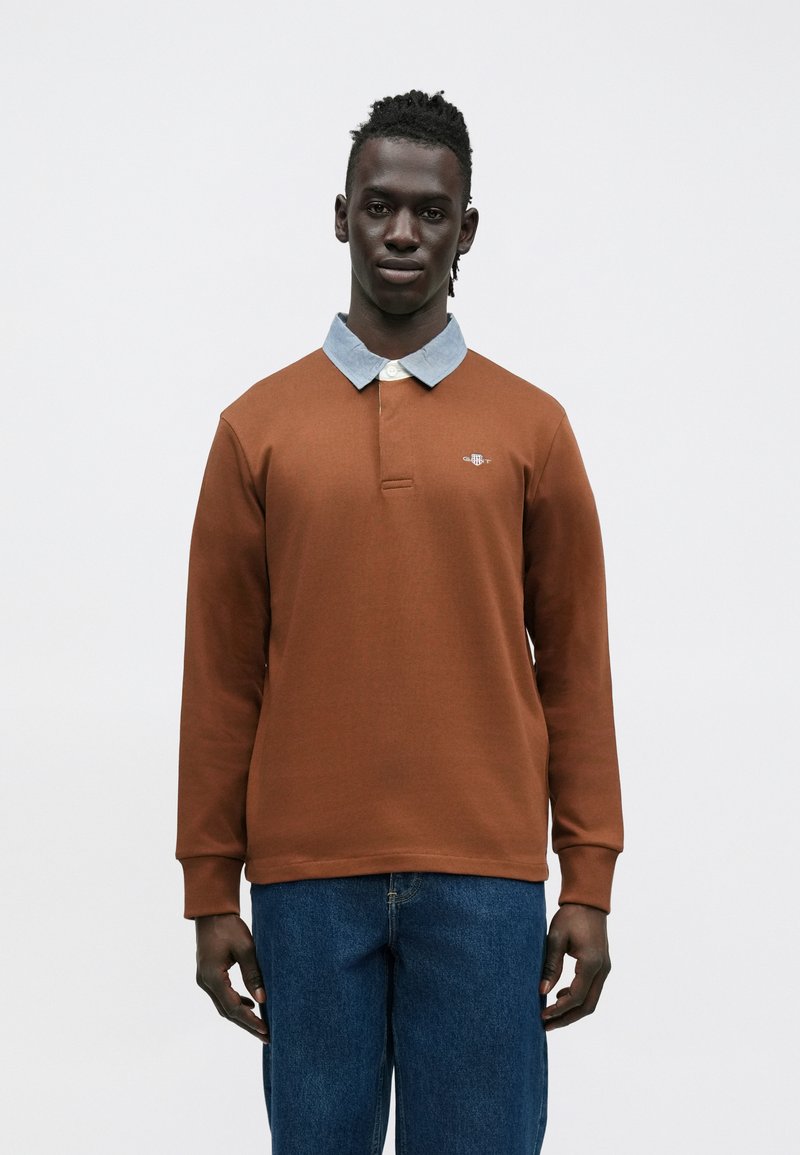 Bruine polo met lange mouwen, lichtblauwe kraag en knoopsluiting, met een klein logo op de borst. Gecombineerd met blauwe spijkerbroek.