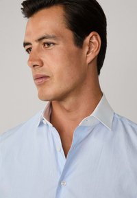 Uomo con i capelli scuri corti che indossa una camicia azzurra chiara con colletto bianco, che guarda a sinistra su uno sfondo semplice.