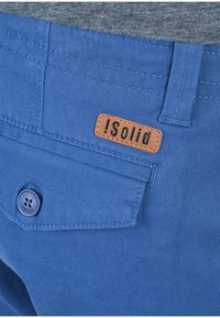Pantaloni blu con una tasca posteriore dotata di bottone e un piccolo etichetta in pelle marrone con la scritta "!solid" cucita sopra la tasca.