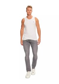 Witte, mouwloze tanktop met een ronde hals gedragen door een model. Grijze slim-fit jeans en witte sneakers maken de outfit compleet.