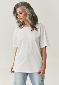 Hvid bomulds t-shirt med korte ærmer, rund halsudskæring og et lille dekorativt mærke ved kanten, parret med lys blå slidte jeans.