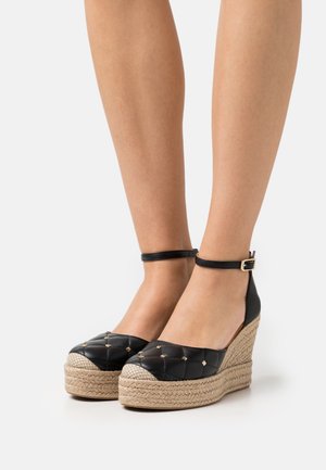 Jambes portant des espadrilles compensées noires matelassées avec des brides à la cheville et des clous dorés sur des talons plateforme tissés sur fond blanc.