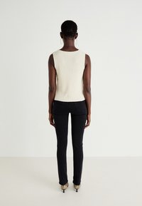 Stickad ärmlös topp i off-white, figurnära design. I kombination med svarta skinny jeans och spetsiga guldklackar, visad bakifrån.