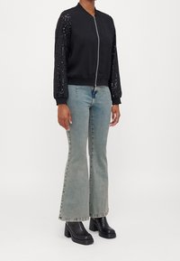 Czarna kurtka bomberka z rękawami ozdobionymi cekinami, ściągaczami przy mankietach i przednim zamkiem błyskawicznym. W połączeniu z jasnoniebieskimi dzwonowymi jeansami i masywnymi czarnymi obcasami.