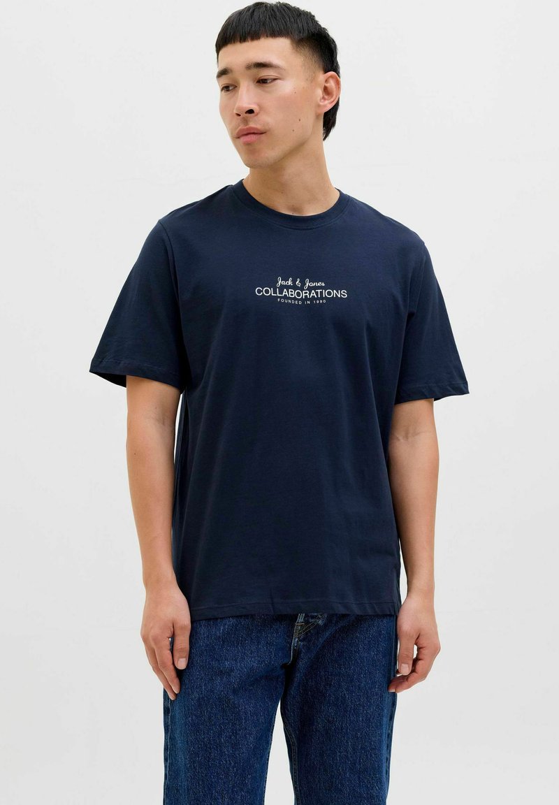 Tumši zila kokvilnas T-kreklu ar īsām piedurknēm, apaļu kakla izgriezumu un baltiem drukātiem uzrakstiem "Jack & Jones COLLABORATIONS FOUNDED IN 1990."