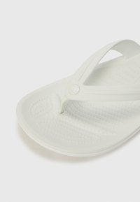Weißer Flip-Flop-Sandal mit einer strukturierten Sohle, Gummiriemen und einem runden Logo auf dem Riemen. Minimalistisches Design mit glatten Kanten.