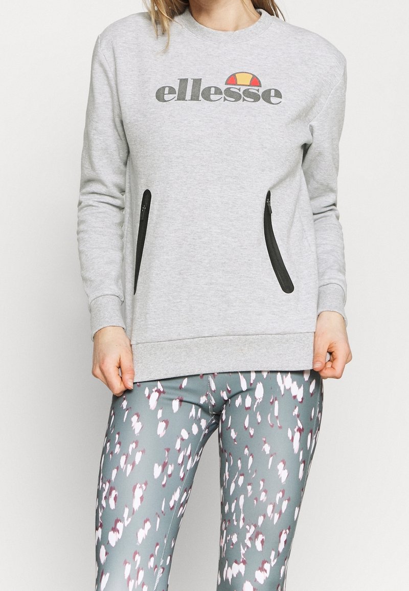 Sweat-shirt gris avec logo, poches zippées sur les côtés, col rond et poignets côtelés. Associé à des leggings à motifs dans des nuances de gris et de blanc.