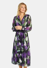 Blumenkleid aus schwarzem, durchscheinendem Stoff, mit langen Ärmeln, einem tiefen V-Ausschnitt und einem Bindegürtel, verziert mit lila und grünen Blumenmustern.