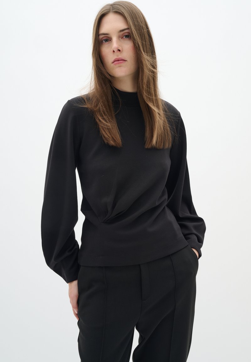 Black long-sleeve top with a high neck, featuring a gathered detail at the waist and smooth fabric, paired with tailored black trousers.