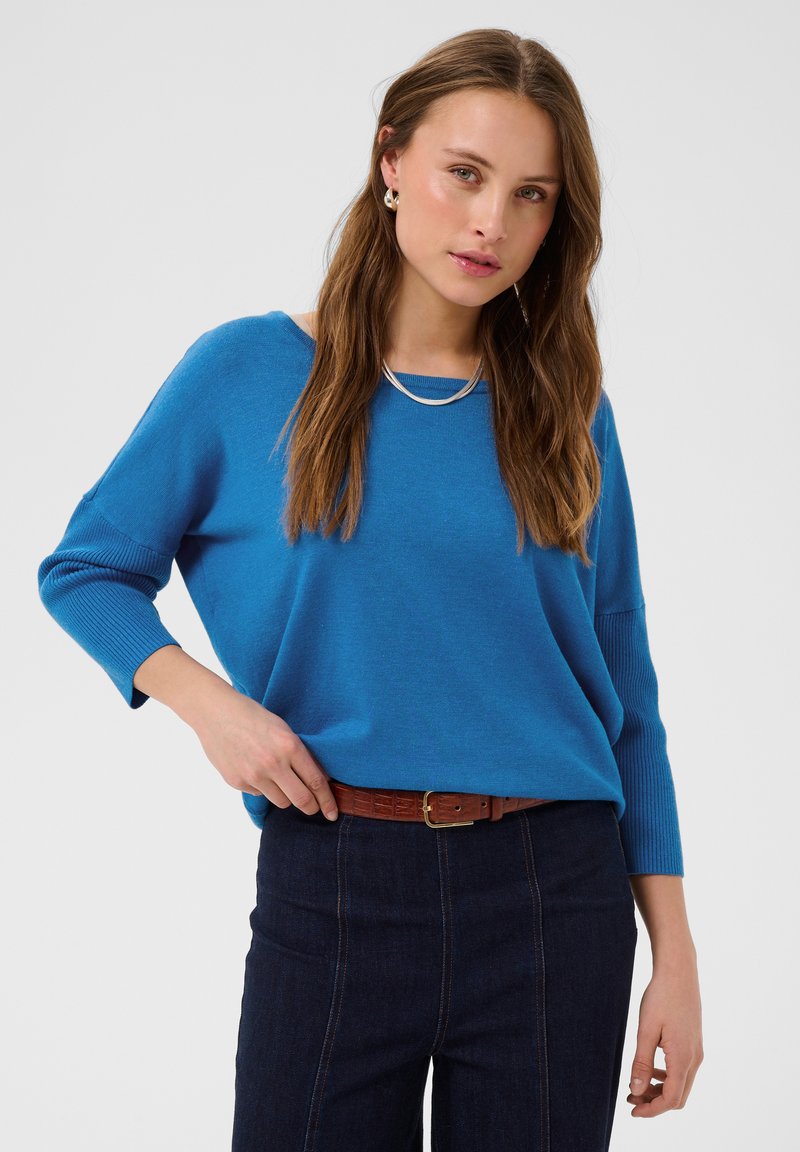 Femme aux longs cheveux bruns portant un pull bleu, un pantalon taille haute foncé, une ceinture marron, un collier en argent et des boucles d’oreilles créoles sur un fond uni.