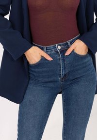 Hoch taillierte Jeans aus dunkelblauem Denim, figurbetonter Stil mit Vordertaschen und Knopfverschluss. Kombiniert mit einem maronenfarbenen, enganliegenden Oberteil.
