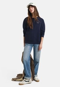 Marineblå hettegenser med rund hals og ribbestrikkede mansjetter, kombinert med lyseblå dressbukser med rette ben og beige sneakers. Rutete stoff ligger i nærheten.