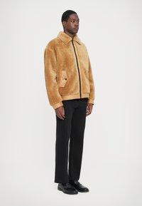 Brun faux fur jakke med lynlås, krave og sidelommer. Kombination med sorte bukser og sko, stående mod en ensfarvet baggrund.