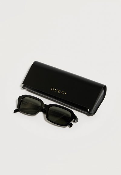 Lunettes de soleil rectangulaires noires avec des verres foncés, posées à côté d'un étui Gucci noir avec un logo doré sur un fond blanc.