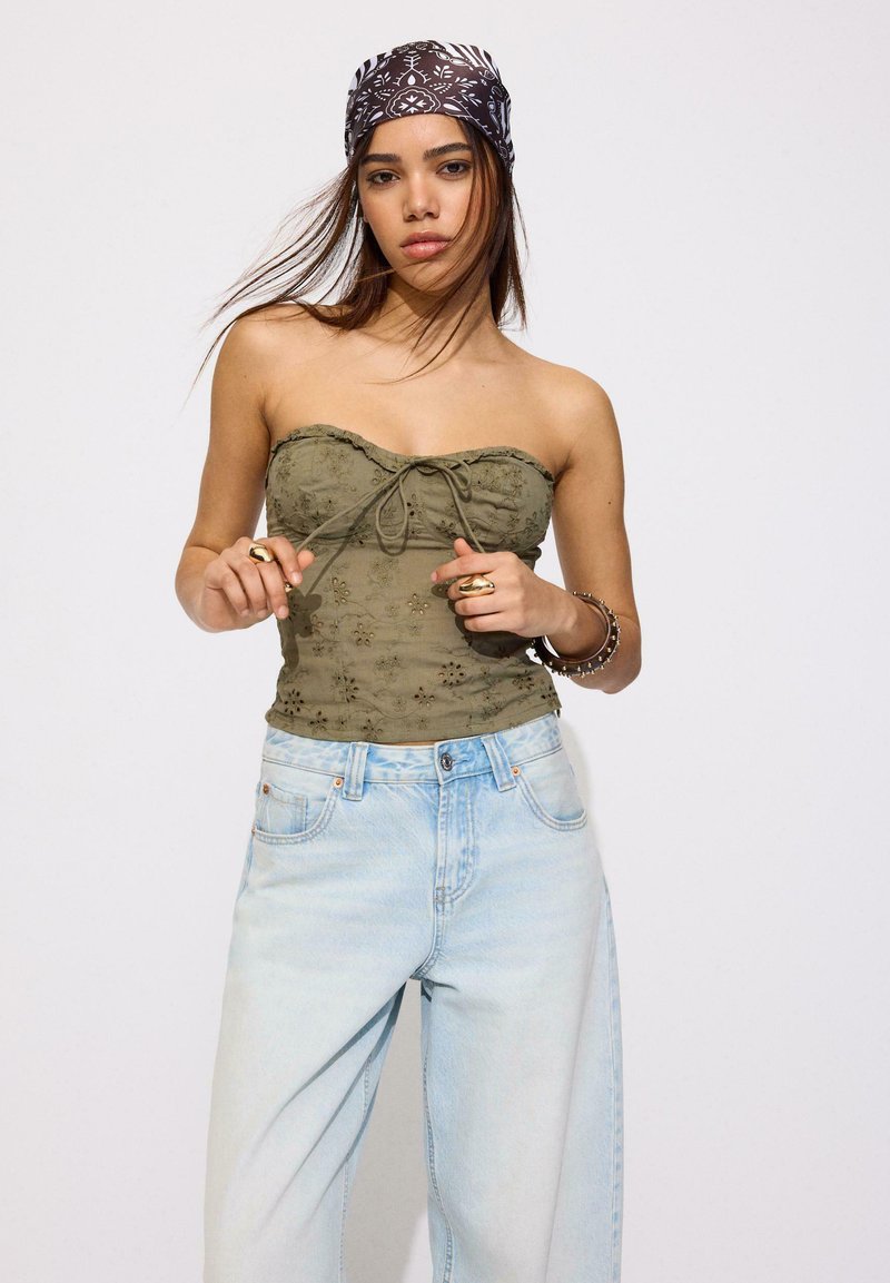 Jeune femme portant un haut bustier vert brodé, un jean bleu clair, un bandana noir à motifs et des bagues, posant devant un fond uni.