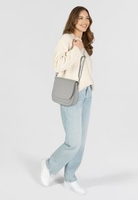 Donna che indossa un maglione leggero e jeans, con una borsa a tracolla grigia, sorridente, in piedi davanti a uno sfondo chiaro e semplice.