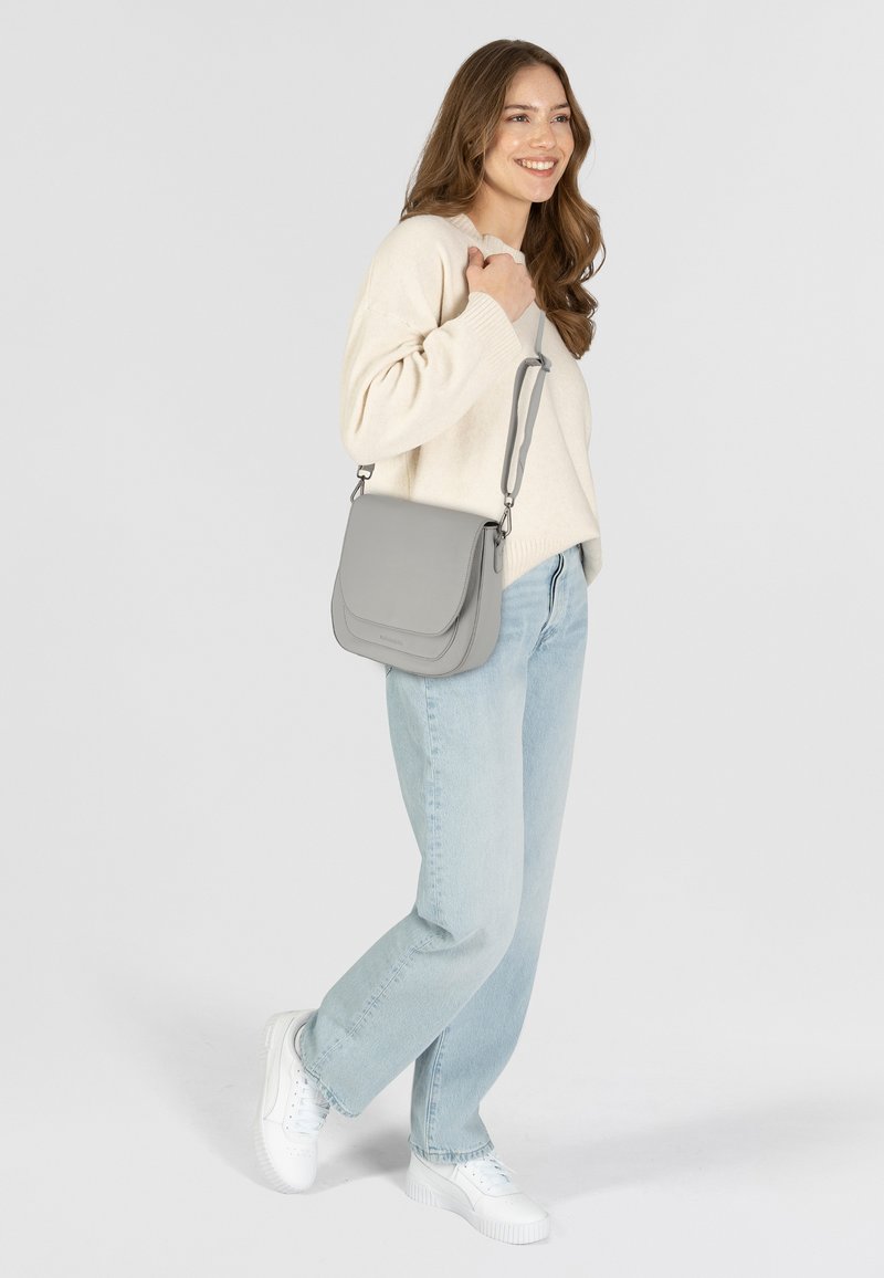 Donna che indossa un maglione leggero e jeans, con una borsa a tracolla grigia, sorridente, in piedi davanti a uno sfondo chiaro e semplice.