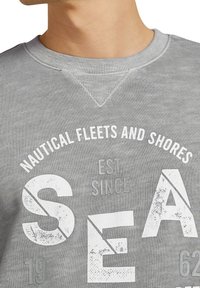 Šedá mikina s žebrovaným výstřihem. Má výrazný, opotřebovaný bílý text s nápisem "NAUTICAL FLEETS AND SHORES" a "SEA" s číselnými akcenty.