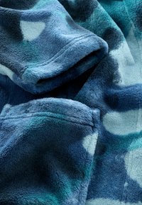 Pătură din fleece în nuanțe de albastru și teal, cu o suprafață moale și texturată și un model abstract. Marginea este cusută pentru durabilitate.