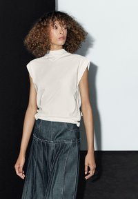 Crème-kleurige mouwloze top met een hoge halslijn, gecombineerd met een donkere denim rok met witte stiksels en plooidetails.