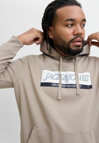 Beige hættebluse med en frontlomme. Har et trykt logo i blå og sort på en hvid baggrund. Blødt stoftekstur.