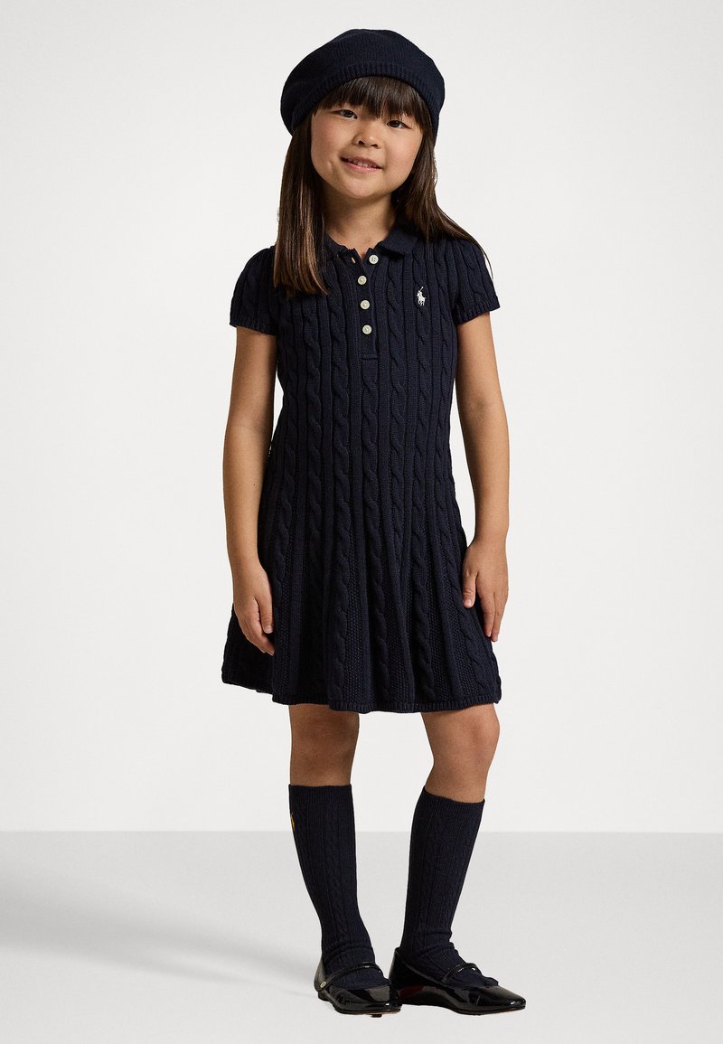 Vestido de punto trenzado en color navy con mangas cortas, con un cuello tipo polo y tres botones, combinado con medias hasta la rodilla y zapatos negros brillantes.
